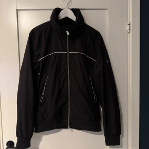 Svart vindjacka från Boomerang - Svart vindjacka från Boomerang med vita detaljer och broderad logga på ärmen. Jackan har hög krage, två fickor med dragkedja och ribbade muddar. Materialet är lätt och syntetiskt, perfekt för blåsiga dagar. Snygg och stilren design med skandinavisk vibe.