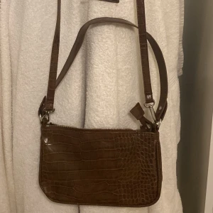 Brun axelväska i krokopräglat fejk skinn - Snygg brun axelväska med krokodilpräglat mönster och justerbar rem. Väskan har dragkedja upptill och ett innerfack med dragkedja. Perfekt storlek för det viktigaste och ger en trendig touch till din outfit☺️