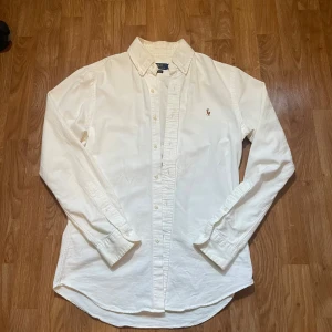 Vit slim fit skjorta från Polo Ralph Lauren - Vit skjorta från Polo Ralph Lauren i slim fit med klassisk krage och knappar hela vägen. Broderad logga med polospelare på bröstet. Tillverkad i mjuk bomull med stretch för extra komfort. Perfekt för dig som gillar stilrena och tidlösa plagg.