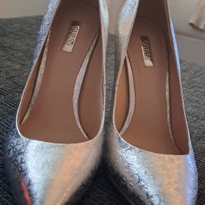 Silvriga pumps från Guess med mönster - Säljer ett par snygga silvriga pumps från Guess med spetsig tå och hög klack. Skorna har ett glansigt material med ett diskret mönster och Guess-logga i metall bak på hälen. Perfekta för dig som gillar att sticka ut med stilrena detaljer.