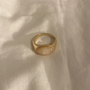 Guldfärgad ring med oval sten - Snygg guldfärgad ring med en oval, ljusbeige sten i mitten. Ringen har en stilren och chunky design som verkligen sticker ut. Perfekt accessoar för att lyfta din outfit med en touch av elegans och trend.