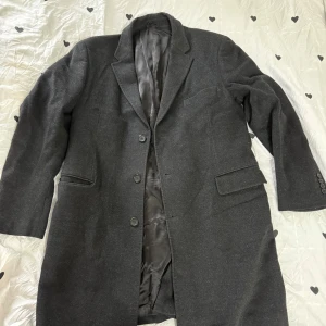 Mörkgrå lång kavaj med knappar - Stilren mörkgrå kavaj med klassisk passform och tre knappar framtill. Kavajen har två fickor med lock och en bröstficka. Fodrad insida och diskreta knappar vid ärmslut. Perfekt för dig som vill ha en tidlös look.