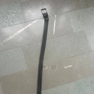 Svart skärp från John Spencer - Snyggt svart bälte från John Spencer med klassisk silverfärgad metallspänne. Bältet är smalt och har stilrena sömmar längs kanterna. Perfekt för att ge din outfit en clean och tidlös touch. Materialet är äkta skinn och spännet har en diskret gravering med märket.