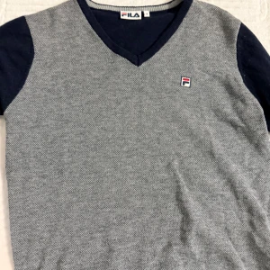 Grå och marinblå stickad tröja FILA - Snygg stickad tröja från FILA i grått med marinblå ärmar och v-ringning. Liten broderad FILA-logga på bröstet. Perfekt för dig som gillar sportig och stilren look. Skön passform och klassisk design.
