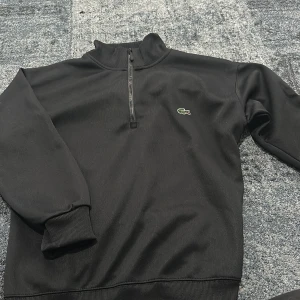 Svart half zip tröja från Lacoste - Svart långärmad tröja från Lacoste med klassisk krokodillogga på bröstet. Tröjan har en hög krage och half zip-dragkedja framtill. Tillverkad i mjukt material med ribbade muddar vid ärmslut och nederkant. Perfekt för en clean och sportig look.