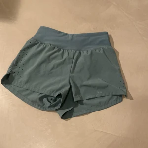 Grågröna träningsshorts från H&M Move - Snygga grågröna träningsshorts från H&M Move med hög midja och bred resår. Shortsen har diskreta håldetaljer på sidorna för ventilation, snörning i midjan och en liten dragkedjeficka bak. Perfekta för löpning eller gymmet.