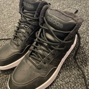 Svarta adidas vinterskor - Säljer ett par svarta adidas sneakers med högt skaft och vit sula. Insidan är fodrad med mjuk päls för extra värme och komfort. Skorna har snörning och tydlig adidas-logga på plösen och hälen. Perfekta för dig som vill ha streetstyle med cozy vibe.