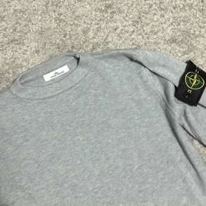 Stone Island Tröja  - Snygg grå stickad tröja från Stone Island i storlek S med klassisk rund hals och ribbade muddar. Tröjan har lång ärm och den ikoniska Stone Island-loggan på vänster ärm. Perfekt för dig som gillar stilrena och trendiga plagg.