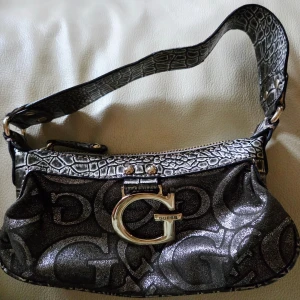 Svart glittrig axelväska från Guess - Cool svart axelväska från Guess med glittrigt material och stora G-loggor över hela väskan. Väskan har ett brett axelband med krokodilpräglat mönster och en stor metallfärgad G-detalj framtill. Perfekt för dig som vill sticka ut.