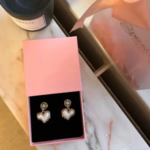 Hjärtformade pärlörhängen i guld - Supersöta örhängen med hjärtformade hängen i pärlvit och guldfärgad kant. Ovanför hjärtat sitter en liten blomliknande detalj i guld. Perfekta för dig som vill ha något gulligt och trendigt i smyckeskrinet.