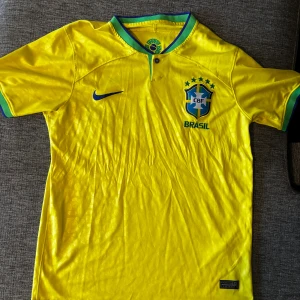 Brasilien Nike fotbollströja gul - Gul Brasilien fotbollströja från Nike med blått och grönt på krage och ärmslut. Broderat CBF-emblem och Nike-logga på bröstet. Tillverkad i Dri-FIT polyester som andas, med subtilt mönster i tyget. Klassisk knapp vid halsen och korta ärmar.