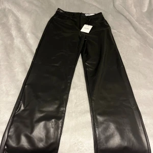 Svarta wide byxor från Vero Moda - Snygga svarta byxor från Vero Moda i wide fit-modell. Byxorna har hög midja och är gjorda i ett glansigt syntetmaterial som ger en cool skinnlook. Klassisk femficksdesign och raka, vida ben. Perfekta för dig som vill sticka ut med en trendig stil.