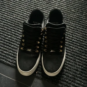 NUBIKK 46 - Svarta sneakers från Nubikk med snygga guldfärgade metalldetaljer vid snörningen och vit platt sula. Skorna har rund tå och är tillverkade i skinn med en stilren och modern look. Perfekta för dig som gillar streetstyle och vill ha något som sticker ut.