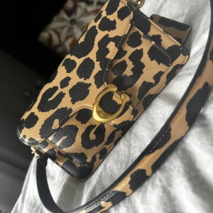 Coach Tabby 20 - Snygg axelväska från Coach i leopardmönstrat skinn med beige och svarta toner. Väskan har en stilren rektangulär form, guldfärgade metalldetaljer och ett praktiskt axelband. Perfekt för dig som vill sticka ut med ett djuriskt statement.