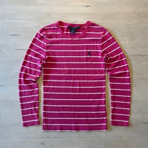 Randig rosa tröja från Ralph Lauren - Säljer en långärmad rosa tröja från Ralph Lauren Sport med vita horisontella ränder och klassisk broderad logga på bröstet. Tröjan är i 100% bomull och har en rund halsringning. Perfekt för dig som gillar preppy och stilren look. Storlek S och i fint skick.