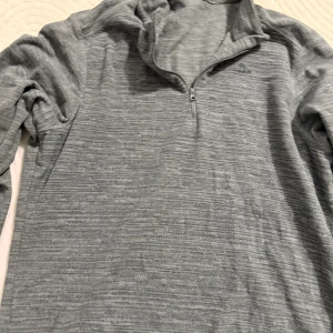 Grå Adidas half zip tröja - Säljer en grå långärmad tröja från Adidas med diskret mönster och half zip-dragkedja framtill. Perfekt för lager-på-lager och chill dagar. Adidas-loggan sitter broderad på bröstet. Skön passform och sportig look.