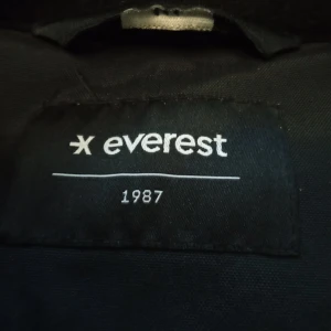 Svart parkajacka med fuskpäls från Everest - Svart parkajacka från Everest med fuskpäls på huvan. Jackan har dragkedja och knappar framtill, samt praktiska fickor med lock. Materialet är slitstark polyester och jackan är perfekt för kalla vinterdagar. Snygg och funktionell design som håller dig varm.