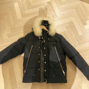 Svart pufferjacka med päls från Horspist - Svart pufferjacka från Horspist Paris med fluffig vit päls på kragen, stora snedställda dragkedjor och tryckknappar framtill. Jackan har quiltad design och detaljer i metall, perfekt för kalla dagar. Insidan har snyggt mönstrat foder och kedjedetalj.