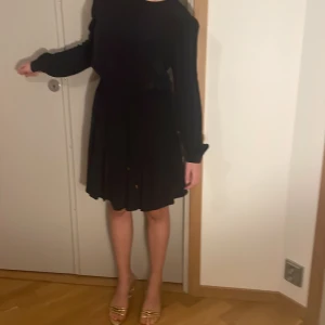 Mörkblå kortklänning från Tommy Hilfiger - Mörkblå klänning från Tommy Hilfiger med långärmade puffärmar och rund hals. Kjolen är lätt plisserad och slutar strax ovanför knäna. Klänningen har en snygg detalj med knappöppning i nacken och är tillverkad i viskos för ett mjukt fall.