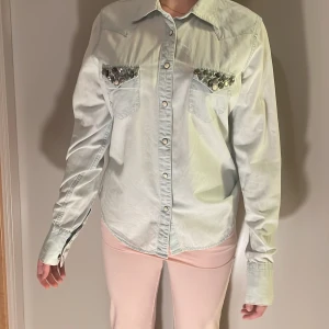 Ljusblå skjorta Ralph Lauren med pärldetaljer - Ljusblå skjorta från Denim & Supply Ralph Lauren med snygga pärl- och metallapplikationer på bröstfickorna. Klassisk westernkrage, tryckknappar och lång ärm. Skjortan är i bomull och har en avslappnad passform.