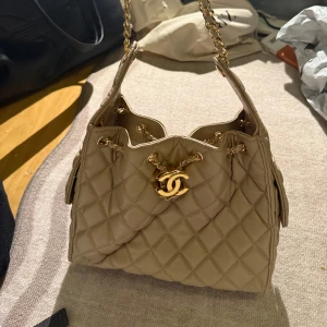Beige quiltad Chanel handväska i skinn - Superfin beige handväska i quiltat skinn med guldfärgade kedjedetaljer och ikoniska CC-loggan framtill. Väskan har snörning upptill och praktiska sidofickor med lock och kedja. Perfekt för dig som vill ha en lyxig och tidlös accessoar. Inte äkta 