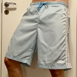 Retro Ljusblå Adidas shorts med snörning - Snygga ljusblå shorts från Adidas med klassiska vita ränder längs sidorna och logga framtill. De har midja med blå snörning och är tillverkade i ett lätt glansigt material. Passformen är loose och längden går till knäna. Märkt M men mer som L