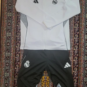Vit Adidas Real Madrid hoodie - Säljer en vit hoodie från Adidas med Real Madrid-logga på bröstet och svarta detaljer. Tröjan har dragkedja framtill, sidofickor och svarta ränder på axeln. Materialet är polyester och passformen är normal. Perfekt för träning eller chill.