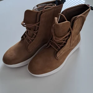 Chunky boots från Tommy Hilfiger - Chunky hybrid boots från Tommy Hilfiger i beige mocka med vit och brun platt sula. Snygg snörning framtill och klassisk Hilfiger-detalj baktill. Perfekt för dig som gillar stilrena och moderna boots med en twist.