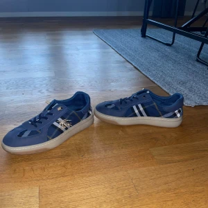 Unika snygga skor från Burberry - Snygga sneakers från Burberry i marinblått med klassiskt rutigt mönster i blått och vitt. Skorna har snörning, rund tå och en platt sula. Materialet är en mix av textil och skinn med broderad Burberry-logga på sidan. Perfekt för dig som gillar stilrena och unika detaljer. Sorleken är 40 men passar också 41. Perfekt för dig som vill ha ett par skor man kan styla till allting. Priset är ej hugget i sten