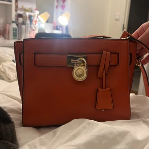 Orange axelväska från Michael Kors - Snygg orange axelväska från Michael Kors i slätt skinn med guldfärgade metalldetaljer och MK-hänglås framtill. Väskan har en fyrkantig form, justerbar axelrem och praktiskt innerfack med dragkedja. Perfekt storlek för det viktigaste och stilren design. Bra som julklapp!