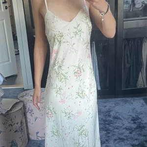 Blommig långklänning från Neo Noir - Superfin långklänning från Neo Noir i ljus, krämig ton med blommigt mönster i rosa och grönt. Klänningen har smala axelband, v-ringad front och öppen rygg med snörning i sidan. Materialet är mjukt och har en satinliknande känsla, perfekt för sommarens alla tillfällen.