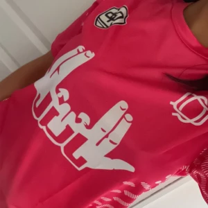 Rosa jul tröja  - Säljer en snygg rosa jul tröja  med vit grafisk print framtill  . Tröjan har korta ärmar och rund hals. Materialet är lätt och andas, perfekt för träning eller match. Unik design med mönstrade detaljer på ärmarna.helt oanvänd, original priset 399kr