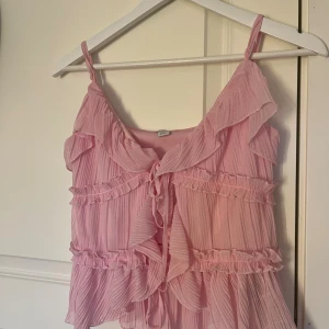 Rosa volangblus från Amisu - Supersöt rosa blus från Amisu med tunna axelband, volanger och knytning framtill. Plisserad och luftig modell som ger en romantisk vibe. Perfekt för dig som gillar en feminin och trendig look. Säljer då den inte kommer till användning, är i mycket bra skick.