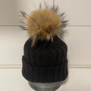 Svart ribbad mössa med pälsboll - Mysig svart stickad mössa med päls boll av tvättbjörn. Knappt använd och fint skick! 