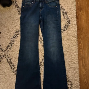 Blå bootcut jeans med låg midja - Snygga blå jeans med bootcut-modell och låg midja. Klassisk femficksdesign och mjukt denimtyg som sitter skönt. Perfekta för dig som gillar en retro vibe och vill ha ett par jeans som sticker ut lite extra.