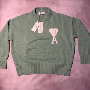 Mintgrön stickad tröja från Ami - Superfin mintgrön stickad tröja från Ami med vit hjärtlogga och A på bröstet. Tröjan har ribbad krage, muddar och nederkant, samt långärmad passform. Perfekt för dig som gillar stilrena detaljer och mjuka material.