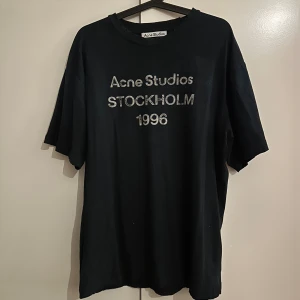 Svart Acne Studios t-shirt - Svart t-shirt från Acne Studios med silverfärgad text 'Acne Studios STOCKHOLM 1996' på bröstet. Modellen är loose fit med korta ärmar och rund hals. Perfekt för dig som gillar stilrena och trendiga plagg med statement-tryck.