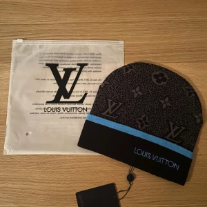 svart mössa med blå kant - Svart mössa med klassiskt monogrammönster och blå kant. Märkets logga är broderad framtill i blått. Snygg och lyxig design som sticker ut, perfekt för dig som gillar exklusiva accessoarer.