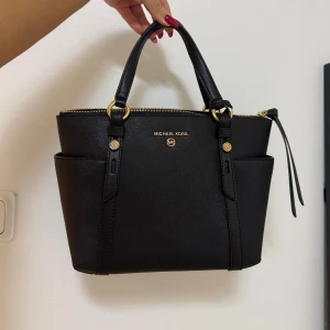 Svart handväska från Michael Kors - Stilren svart handväska från Michael Kors med guldfärgade detaljer och logotyp framtill. Väskan har två handtag, praktiska sidofack och dragkedja upptill. Tillhörande långt axelband finns. Köpt för 3000. Använt 2 gånger