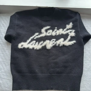 Svart stickad Saint Laurent tröja - Svart stickad tröja från Saint Laurent med vit fluffig logotyp framtill. Tröjan har rund halsringning, ribbade muddar och långa ärmar. Perfekt för dig som vill ha en statement piece med lyxig känsla och cool design.