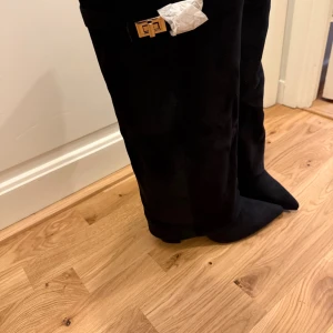 Svarta mockastövlar med spetsig tå - Snygga svarta stövlar i mocka med spetsig tå och låg klack. Stövlarna har en dekorativ guldfärgad spänne upptill och dragkedja på sidan för enkel påtagning. Perfekt för dig som vill ha en stilren och edgy look. Aldrig använda, kmr i orginalförpackning o allt. Pris kan diskuteras🥰