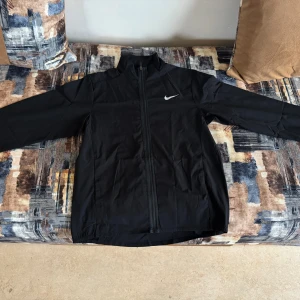 Svart Nike vindjacka Dri-Fit - Svart vindjacka från Nike med Dri-Fit-teknologi, hel dragkedja och ståkrage. Jackan har lång ärm, två sidofickor och en vit Nike-logga på bröstet. Perfekt för dig som gillar sportig och clean stil.
