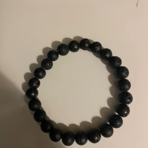 Svart pärlarmband med lava-look - Stilrent svart armband med runda pärlor som har en cool lava-sten struktur. Perfekt för dig som gillar minimalistiska accessoarer med lite edge. Elastiskt band gör det enkelt att ta på och av. Passar både till jeans och hoodie.
