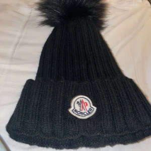 Svart Moncler mössa med pälsboll - Ribbstickad svart mössa från Moncler med stor fluffig pälsboll på toppen och klassisk Moncler-logga framtill. Mössan har uppvikt kant och är tillverkad i mjukt stickat material som håller dig varm och snygg under kalla dagar. OBS! Ej äkta päls, köpt utomlands(ej äkta) för 50 euro!
