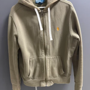 Beige hoodie från Polo Ralph Lauren - Snygg beige hoodie från Polo Ralph Lauren med dragkedja, huva med vita snören och klassisk broderad logga i orange på bröstet. Tillverkad i bomull och polyester, med två fickor framtill och ribbade muddar.skulle säga att denna hoodie sitter som storleken S/M!