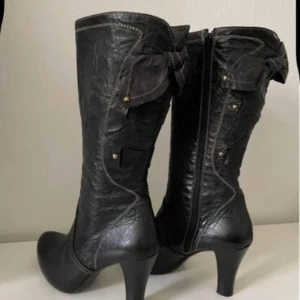 Svarta skinnstövlar med rosett och klack - Snygga svarta stövlar i skinn med hög klack och rund tå. Dekorativa rosetter på sidan och kontrastsömmar ger en cool detalj. Dragkedja på insidan för enkel på- och avtagning. Perfekt för dig som vill ha en edgy look med lite extra attityd. Sömmen släppte , kolla sista bild och en mitt fattas (syns inte)