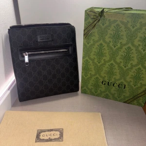 Svart axelväska från Gucci - Snygg svart axelväska från Gucci med klassiskt GG-mönster och silverfärgad dragkedja framtill. Väskan har en fyrkantig form och är tillverkad i slitstark polyester med detaljer i skinn. Perfekt för dig som vill ha en stilren och lyxig väska.
