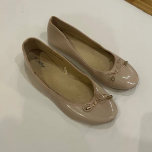 Beige/Rosa ballerinaskor från Alley - Snygga beige/Rosa ballerinaskor från Alley med rund tå och platt sula. Skorna har en glansig finish och en söt rosettdetalj framtill med guldfärgade metalländar. 