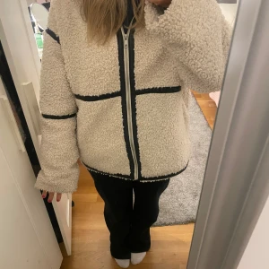 Beige teddyjacka med svarta detaljer - Mysig beige teddyjacka med svarta kantband och dragkedja framtill. Jackan har en loose passform och långärmade ärmar. Perfekt för dig som gillar chill och streetwear-vibe. Materialet är fluffigt och ger en skön känsla under höst och vinter.