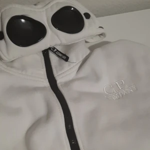 Vit jacka från C.P. Company - Säljer en vit gogglejacka från C.P. Company med ikoniska svarta glasögon i huvan och broderad logga på bröstet. Jackan har hel dragkedja framtill och är tillverkad i mjukt material. Perfekt för dig som vill sticka ut med en unik och stilren look.